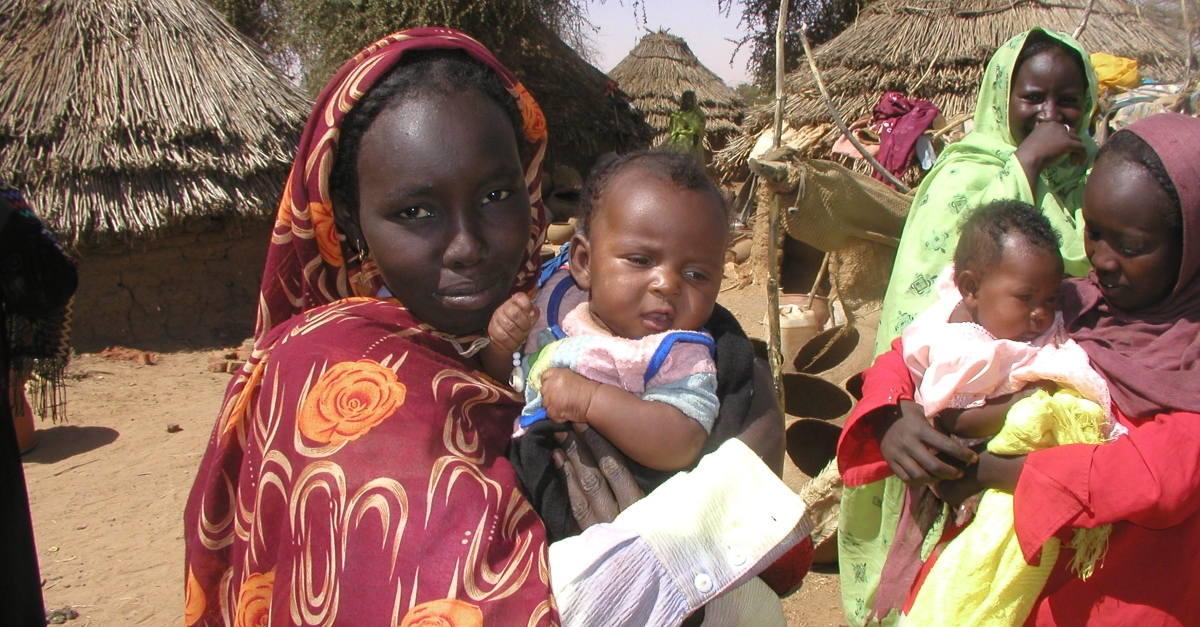 A woman holds a baby in the Darfur region of Sudan. Other women with children and mud structures are in the background.